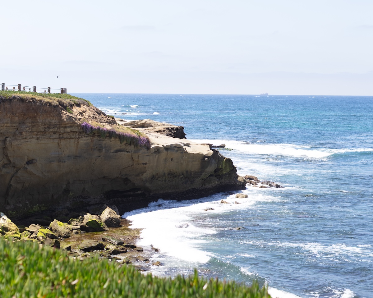 2G2A3108 Waves crashing along rugged coastal cliffs beneath a clear blue sky.